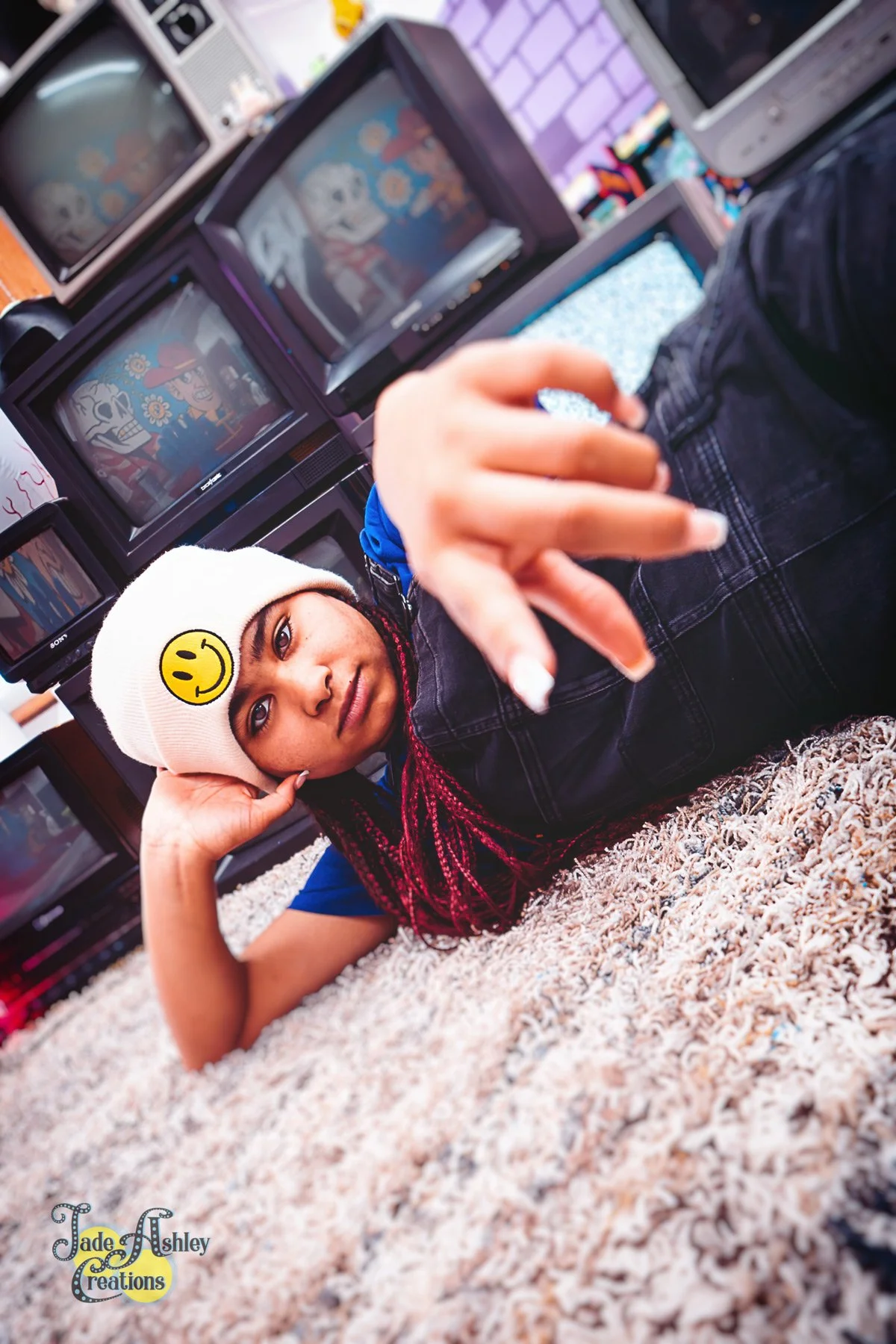 A young woman with pink hair, lying on a carpeted floor, wearing a white beanie with a yellow smiley face, pointing at the camera, with multiple vintage televisions behind her displaying cartoon characters.