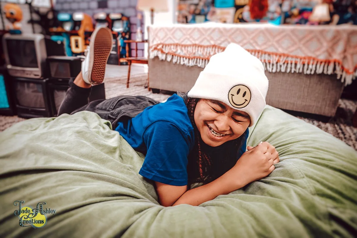 A young boy with a big smile, wearing a white beanie with a smiley face sticker, lying on his stomach on a large green bean bag chair, in a home setting with vintage TVs, a couch, and a table in the background.