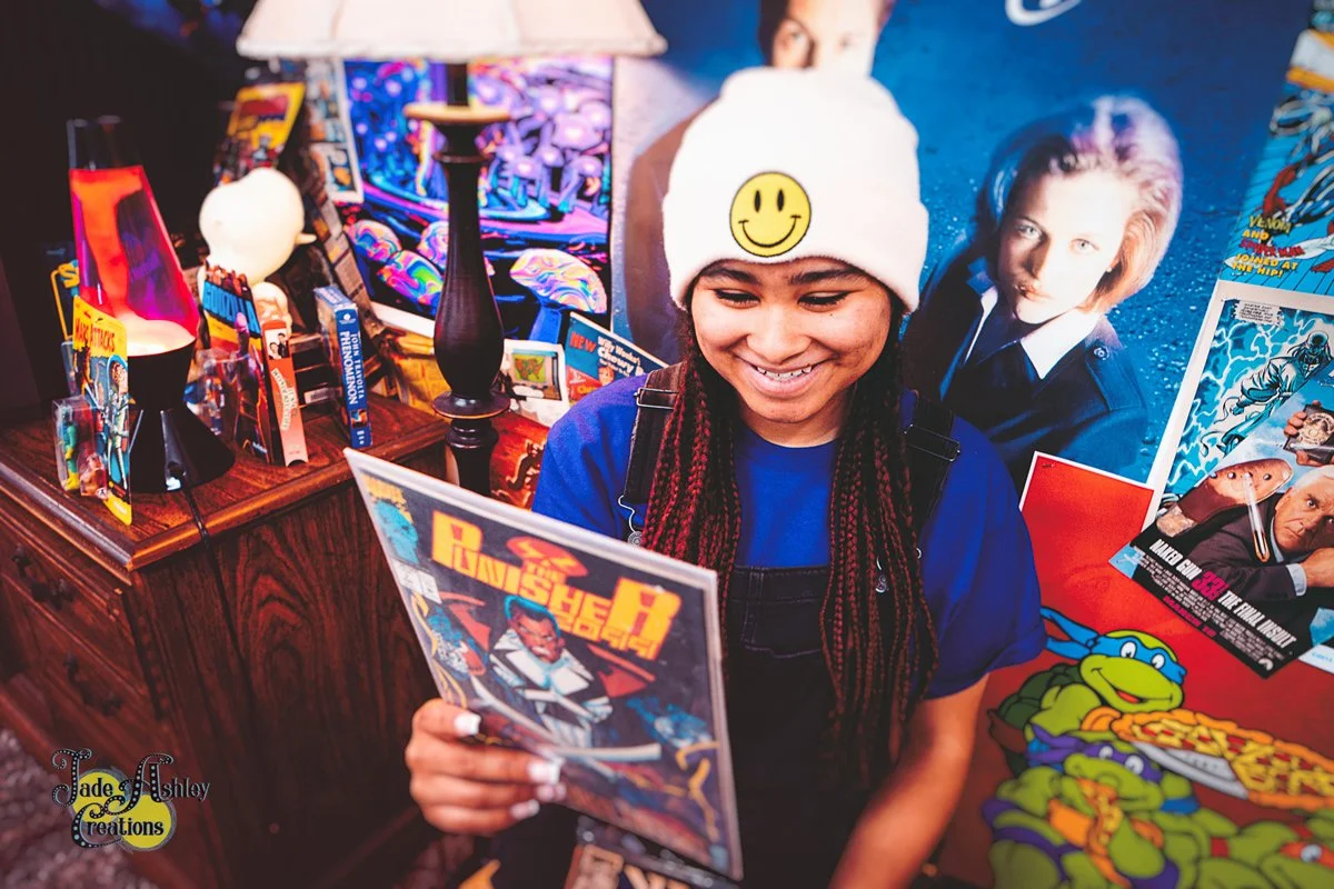 A smiling girl holding a comic book in front of a wall decorated with posters of comic book characters, including Teenage Mutant Ninja Turtles, and vintage comic books on a wooden table.