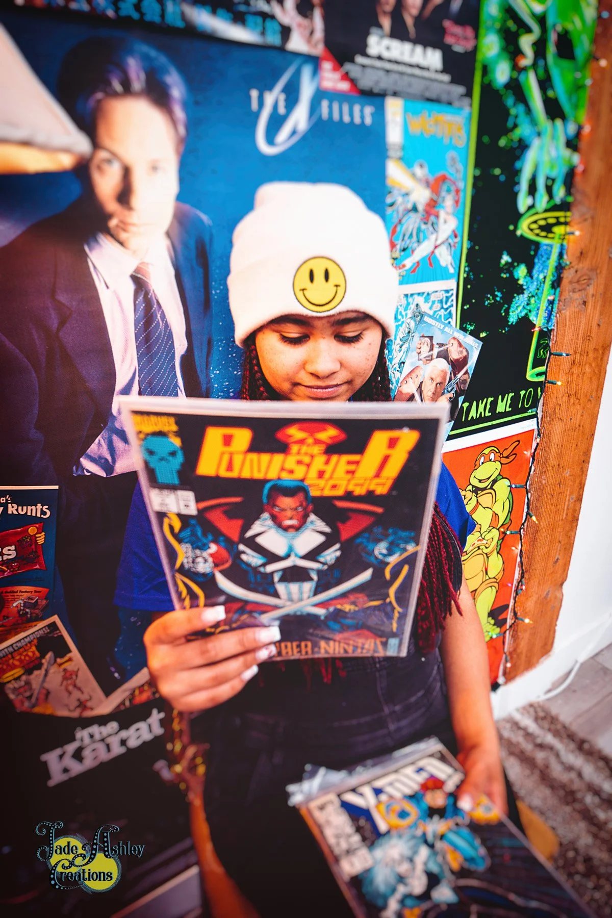 A young girl with braided hair, wearing a white beanie with a smiley face, is reading a comic book titled 'The Punisher.' She is standing in front of a colorful wall decorated with various posters and artwork, including a large poster of a man in a s