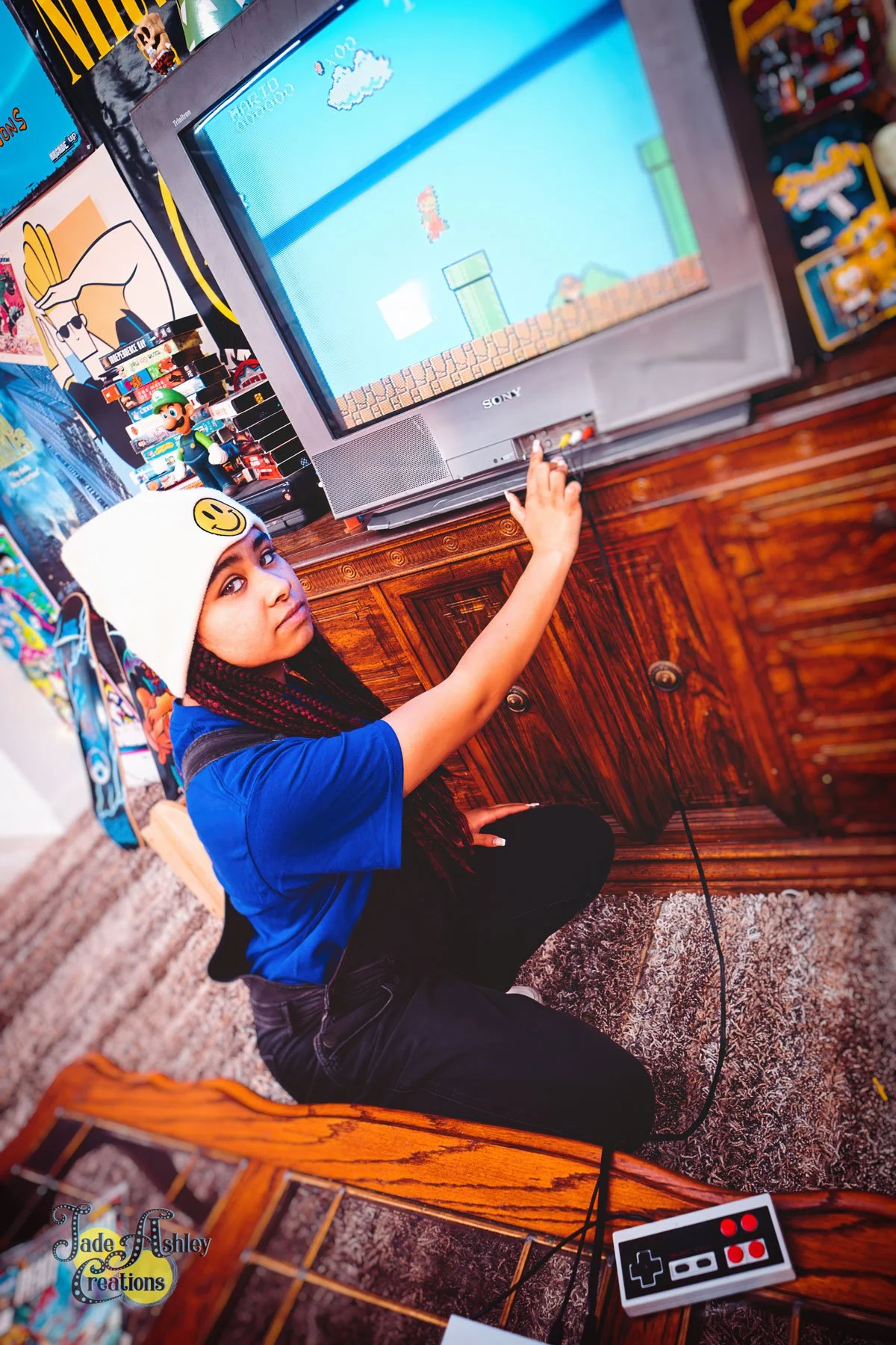 A young girl in a white beanie with a smiley face, wearing a blue shirt, sitting on a carpeted floor, playing a classic video game on a CRT television. She is using a vintage game controller with a cord, and the screen shows a Super Mario game.