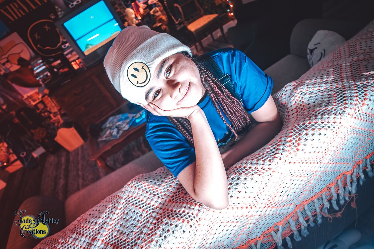 A young girl with braided hair wearing a beanie hat with a smiley face sticker, resting her head on her hand and smiling at the camera in a cozy, dimly lit indoor setting.