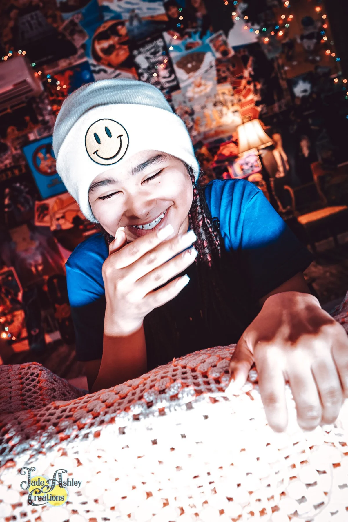 A woman wearing a beanie with a smiley face and a blue shirt, laughing and covering her mouth, sitting at a table with a crocheted tablecloth. The background is decorated with eclectic posters and string lights, creating a cozy, colorful atmosphere.