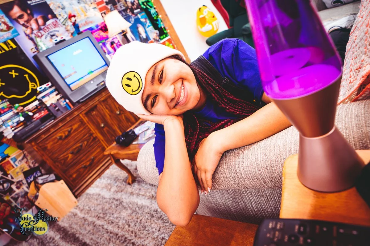 A smiling young woman with braided hair and a white beanie with a yellow smiley face sticker, lying on a beige couch with her head resting on her hand in a colorful, cluttered room. There is a lava lamp on a wooden table, a vintage TV, and a collage 