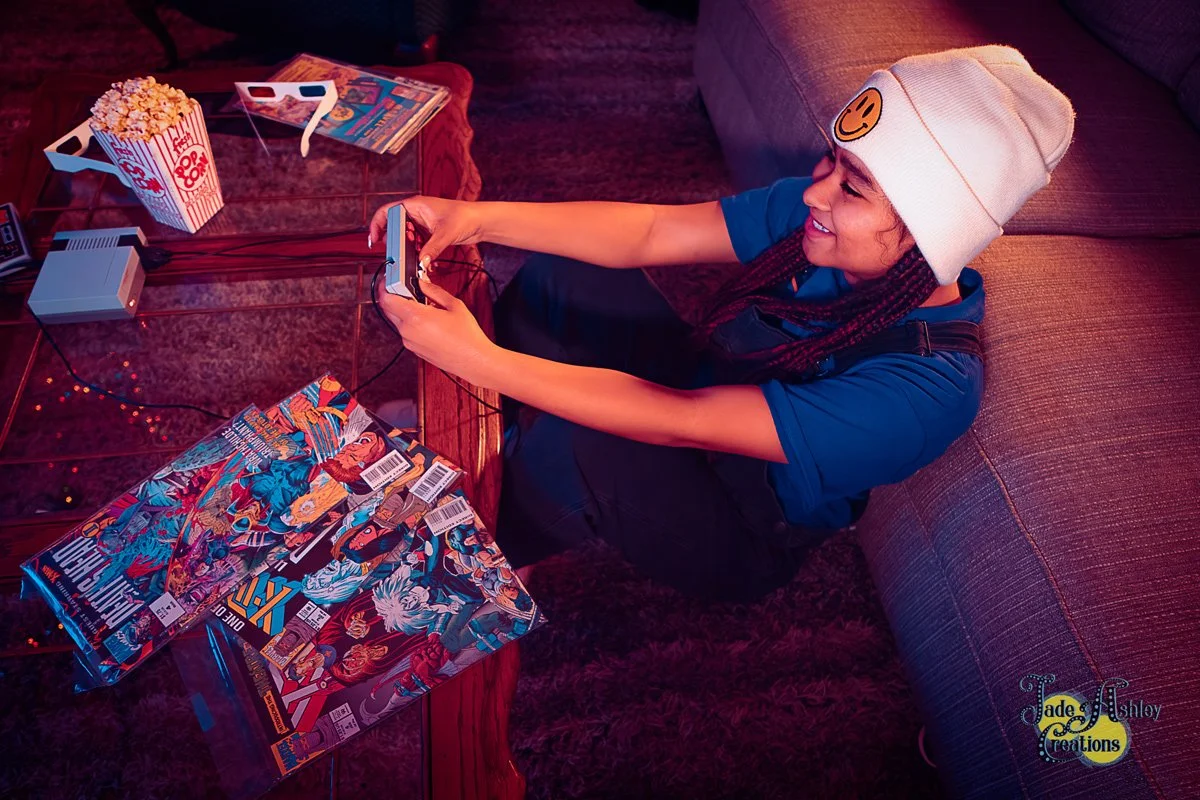 A young girl sits on the floor in a living room, smiling while playing a video game on her phone. She wears a white beanie with a smiley face and a blue shirt. A box of popcorn and comic books are on the glass table in front of her.