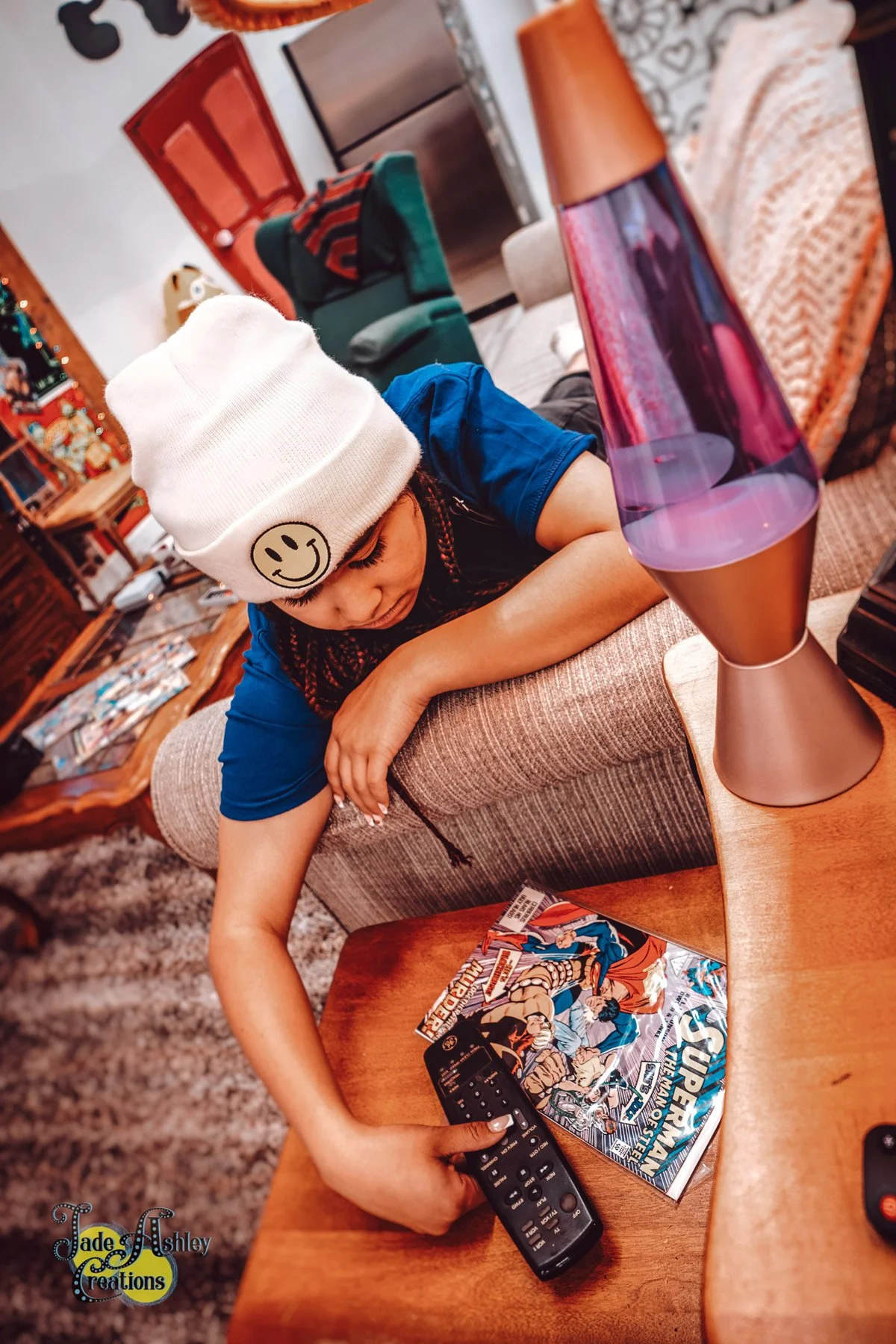 A young girl wearing a white beanie with a smiley face patch, lying on a couch, reaching for a TV remote. There is a lava lamp, comic books, and a remote control on a wooden table nearby in a cozy living room.