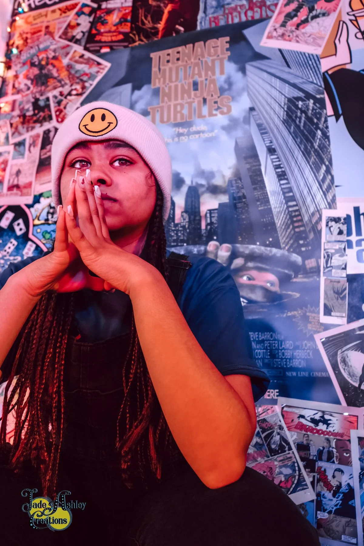 A young woman wearing a white beanie with a smiley face patch, with hands clasped near her face, posing in front of a multicolored collage and a 'Teenage Mutant Ninja Turtles' poster.