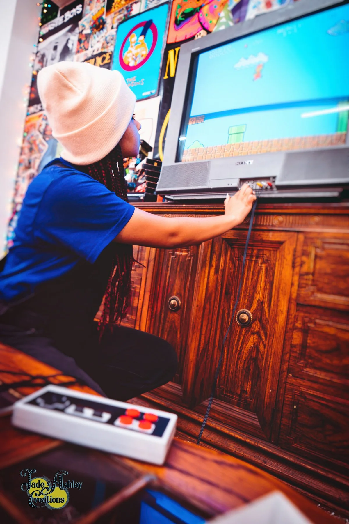A young person wearing a beige beanie and blue t-shirt playing a video game on a flat-screen TV. The person is sitting on a wooden cabinet, with their right hand on a video game console controller. The background has colorful posters, including a blu