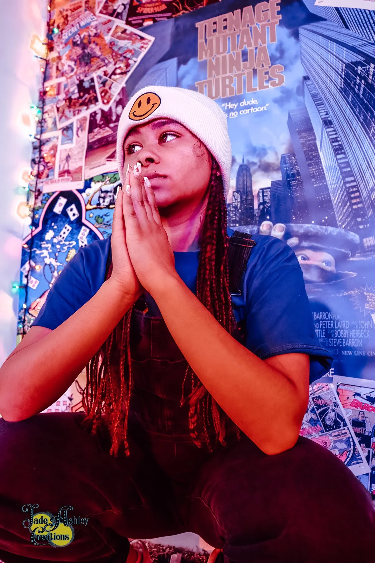 A young girl with braided hair and a white beanie with a smiley face patch, sitting with her hands together near her face, in a room decorated with colorful posters and string lights, including a large 'Teenage Mutant Ninja Turtles' poster.
