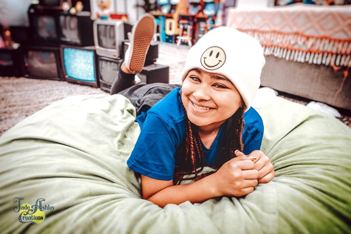 A young girl with braided hair, wearing a white beanie with a smiley face, lying on her stomach on a green bean bag chair, smiling at the camera in a cluttered living room with old TV sets and books in the background.