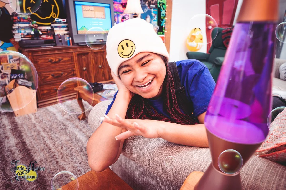 A young girl with braided hair, smiling and resting her head on her hand, looks at a purple lava lamp on a table in a colorful living room. She wears a white beanie with a yellow smiley face.