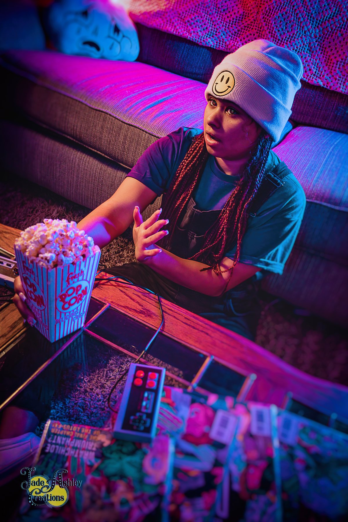 A young woman with braided hair and a beanie with a smiley face, sitting on the floor in a dimly lit, colorful room, watching video game on a TV with a remote in front of her. She has a pained expression and a tear running down her cheek, with a bowl