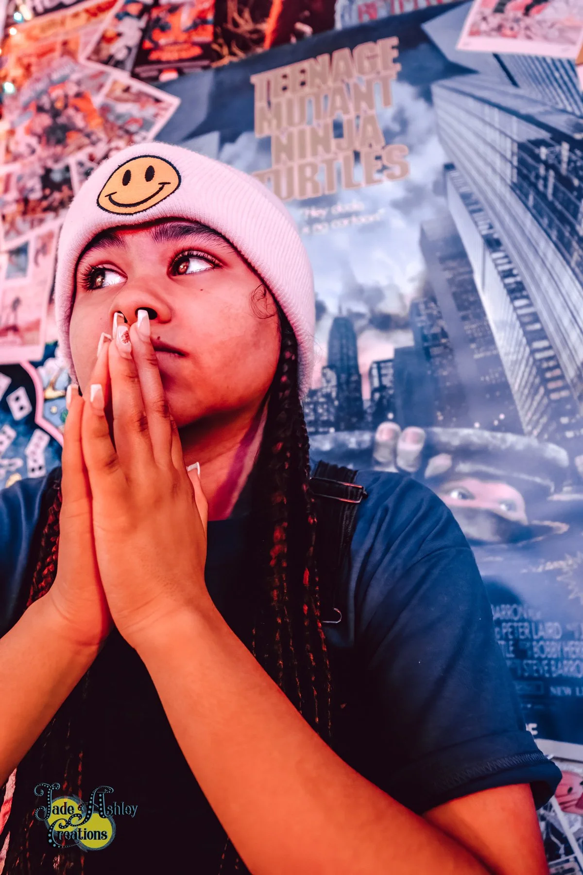 A young person with braided hair is wearing a pink knit cap with a smiley face patch, holding their hands near their face, looking to the side. Behind them is a Teenage Mutant Ninja Turtles poster showing a city skyline.