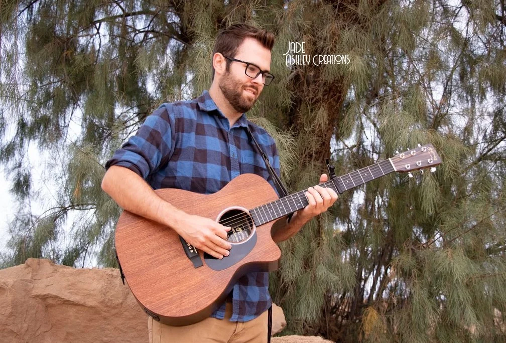 A man wearing glasses and a blue checkered shirt playing an acoustic guitar outdoors, standing in front of trees and rocks.