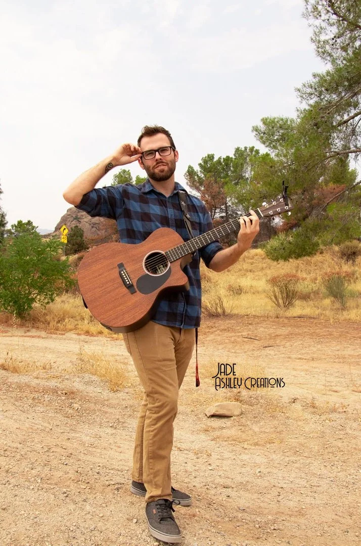 A man standing outdoors in a desert-like area with trees in the background, holding an acoustic guitar, wearing glasses, a blue plaid shirt, khaki pants, and black shoes.