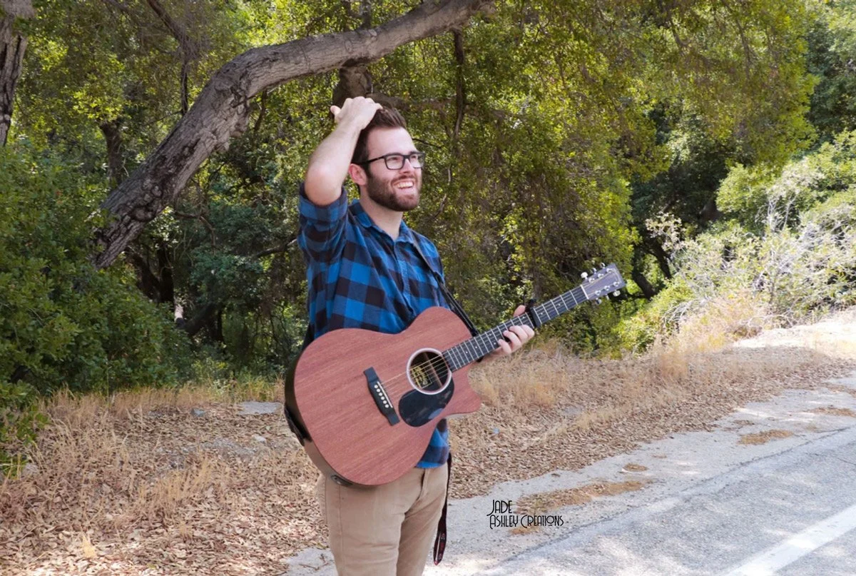 A young man with glasses, a beard, and a blue plaid shirt, standing outdoors on a dirt path surrounded by green trees, holding an acoustic guitar and smiling.