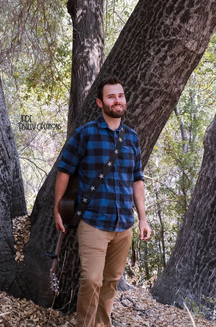 A young man with a beard and brown hair smiling in a wooded area, wearing a blue and black plaid shirt and khaki pants. He is holding a guitar and glasses in his left hand, and standing near large trees with sunlight filtering through the leaves.