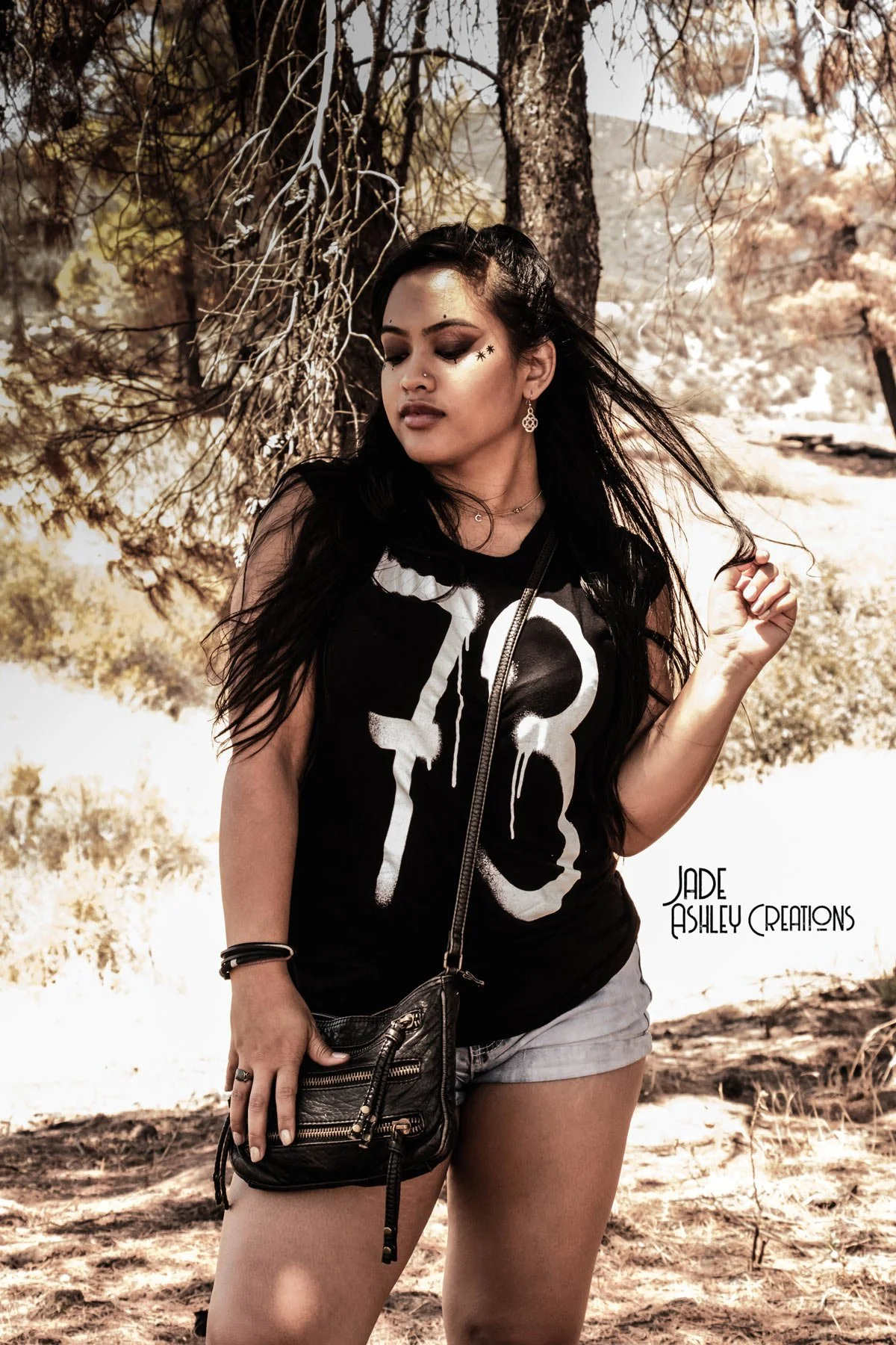 A young woman with long dark hair, makeup, and jewelry stands outdoors with a tree in the background. She is wearing a sleeveless black T-shirt with a large white graphic, denim shorts, and carries a small black crossbody bag. The woman appears to be