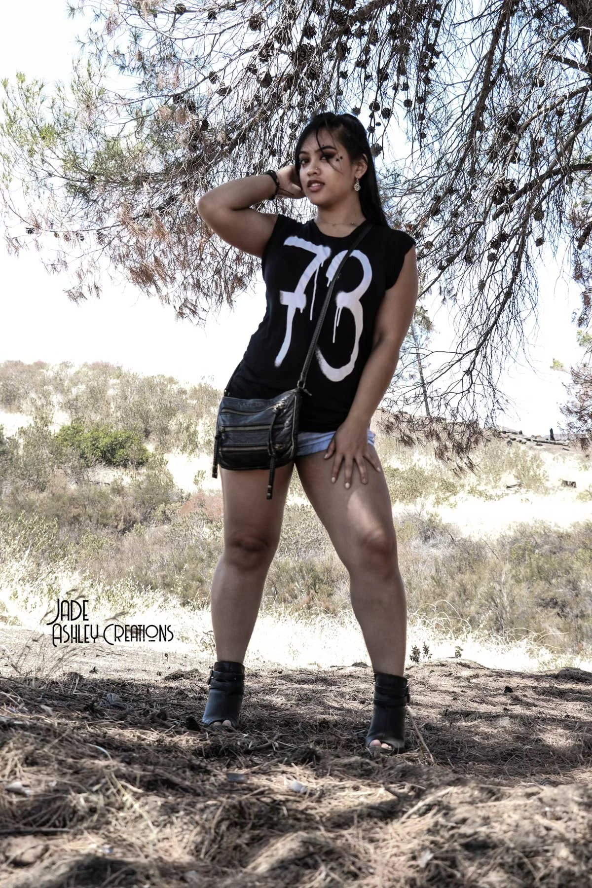 Young woman standing outdoors under a tree, wearing a sleeveless black shirt with white numbers, shorts, and high-heeled boots, holding one hand to her head and the other resting on her thigh, with a small crossbody bag, in a natural landscape.