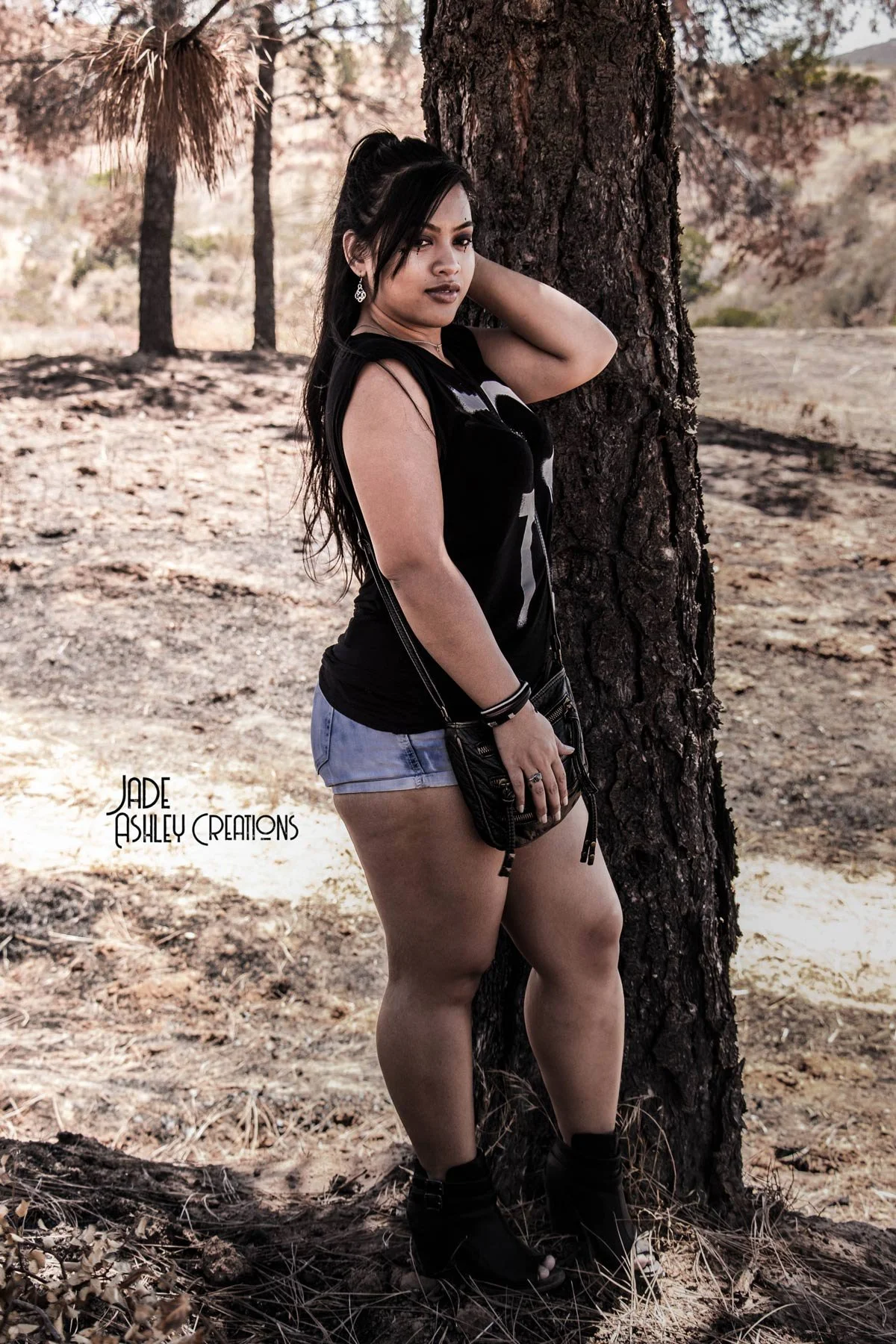 A woman standing outdoors in front of a tree, wearing a black sleeveless top, denim shorts, and dark boots. She has long dark hair and is posing with her hand behind her head, with a handbag hanging from her shoulder.