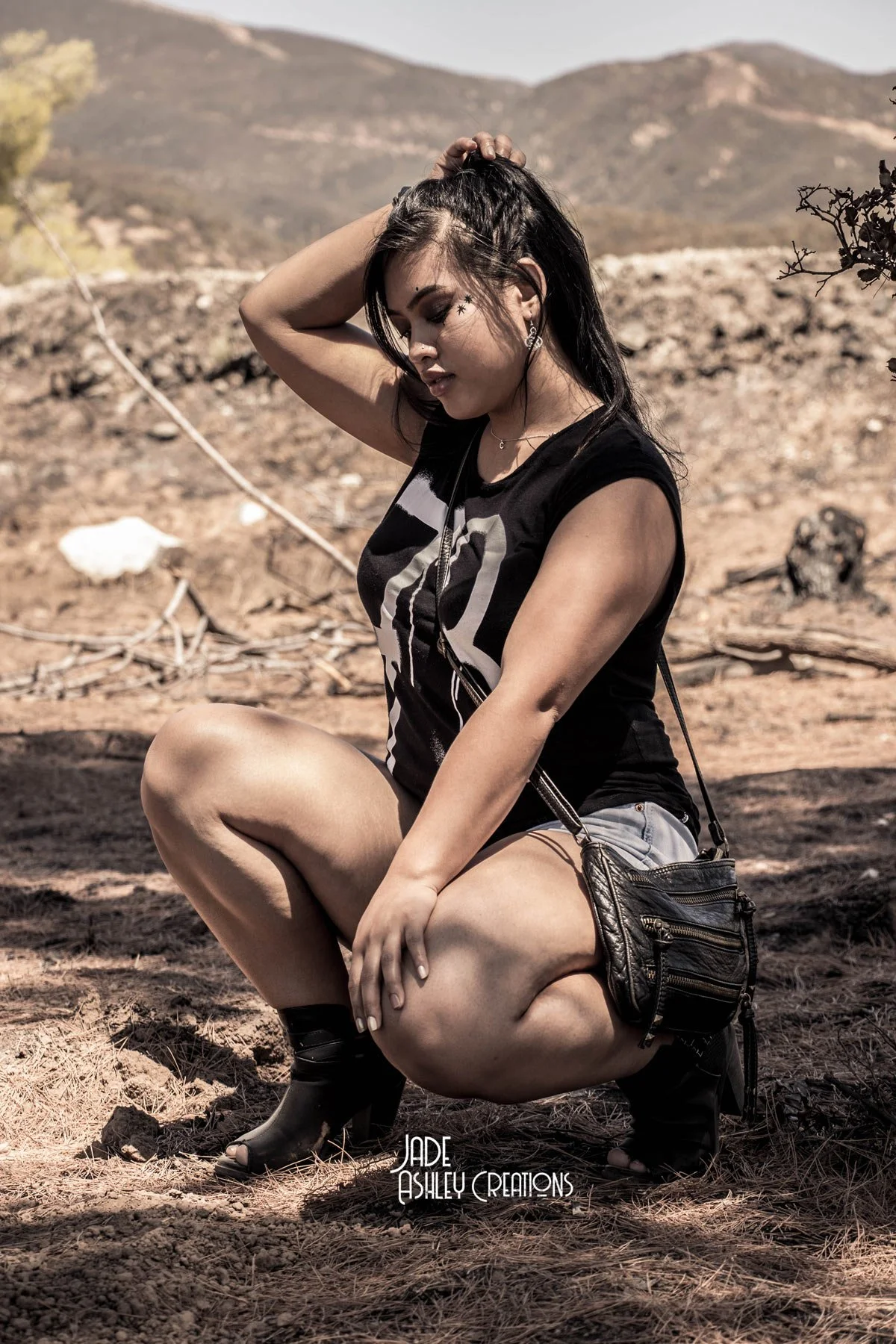 A woman in black top and shorts squatting outdoors on dirt ground with mountains in the background, hand on her head, with dark hair and makeup, wearing earrings, a small necklace, carrying a black purse, and wearing black shoes.
