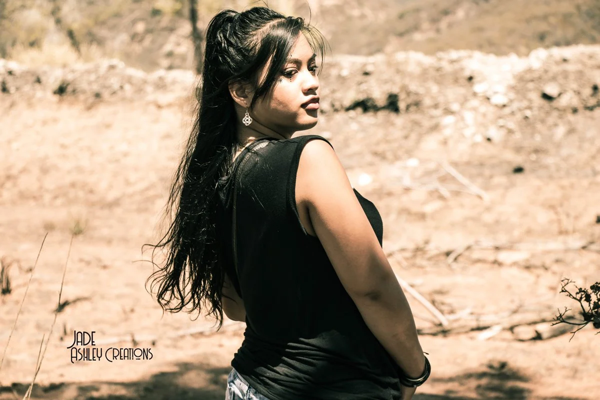 Young woman with long dark hair, wearing earrings, black sleeveless top, standing outdoors in a desert-like area with sandy ground and sparse vegetation.