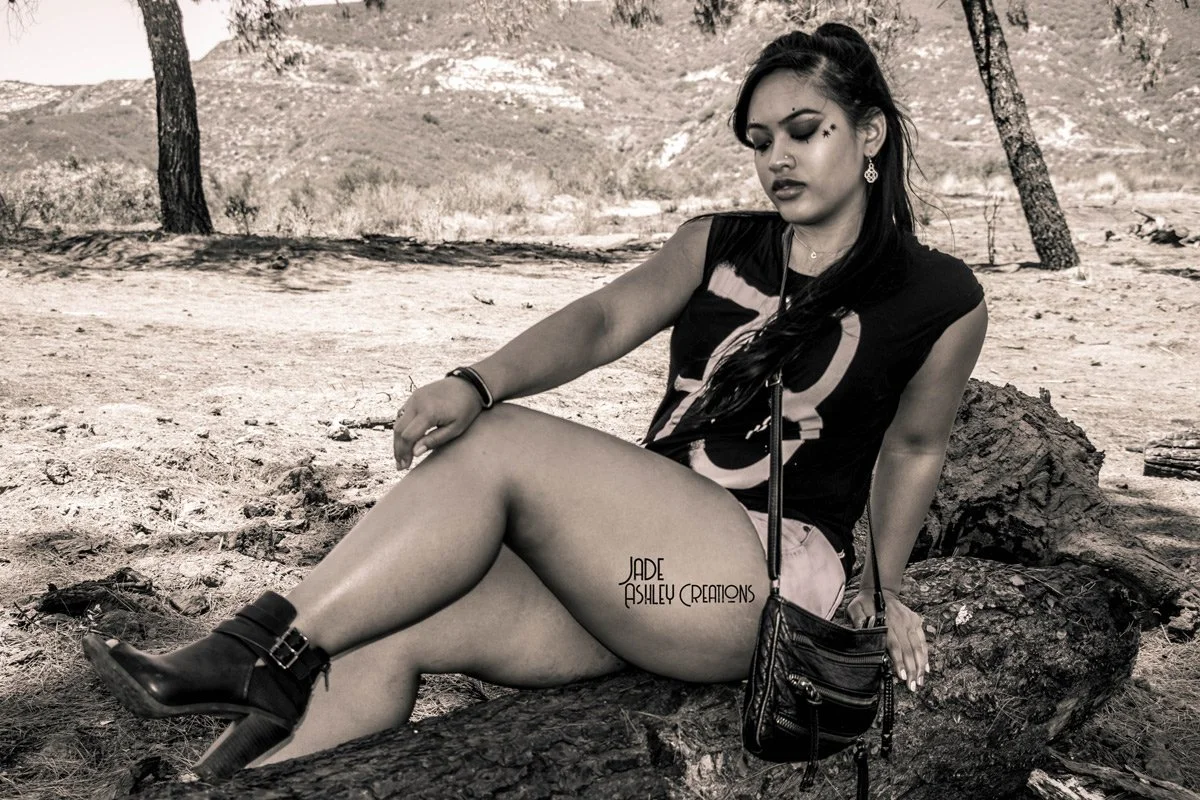 A young woman with dark makeup and earrings sits on a fallen tree trunk in a wooded outdoor area. She is wearing a sleeveless t-shirt, shorts, and ankle boots, and has a handbag resting on the ground next to her. The image is in black and white.