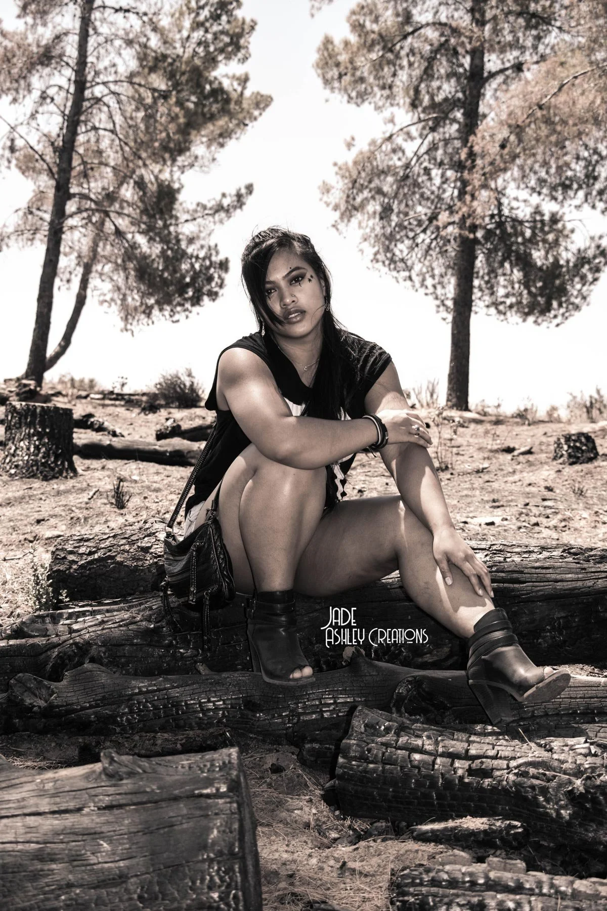 A woman sitting on a log in a forest, with trees and logs around her. She is wearing open-toe boots, black shorts, and a black top, with dark makeup and long hair.