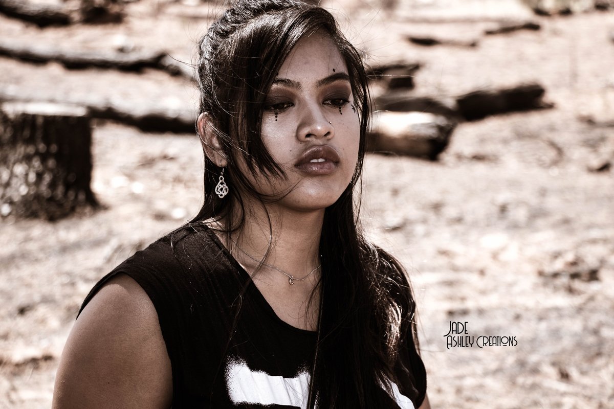 A young woman with long dark hair, wearing makeup and jewelry, standing outdoors on a sandy, rocky terrain with driftwood in the background.