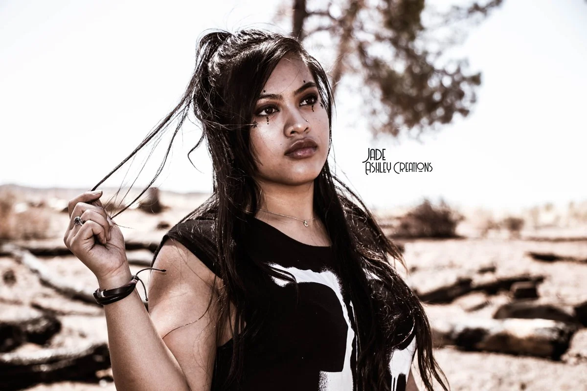 Young woman with long dark hair outdoors in a desert, looking to the side with a serious expression, holding a strand of hair in her hand, wearing a sleeveless black shirt with white print, and jewelry including a necklace and bracelets.