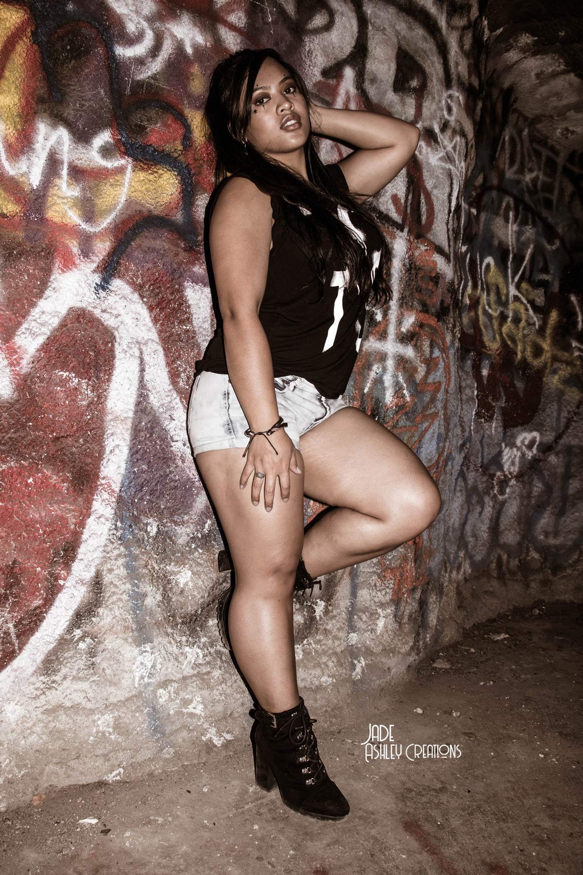 A woman posing against a graffiti-covered wall, wearing a black sleeveless top, white shorts, and black boots, with one foot on the wall and her hand resting on her head.