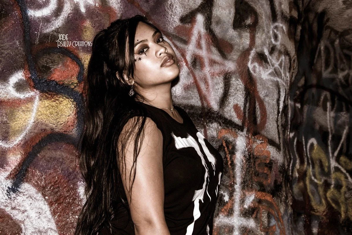 A woman with long dark hair and makeup posing against a graffiti-covered wall.