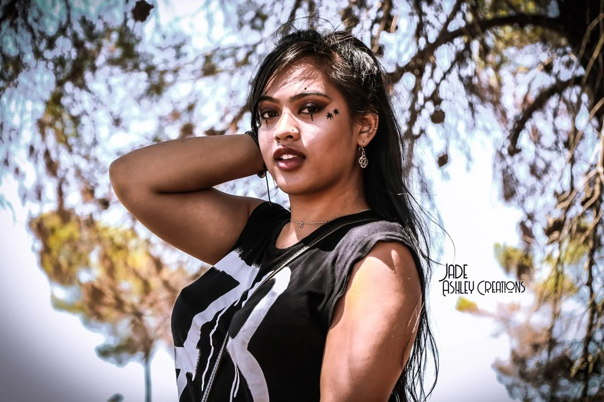 Young woman with dark hair and tattoos, wearing a black sleeveless top, standing outdoors under a tree with an expressive look during daytime.