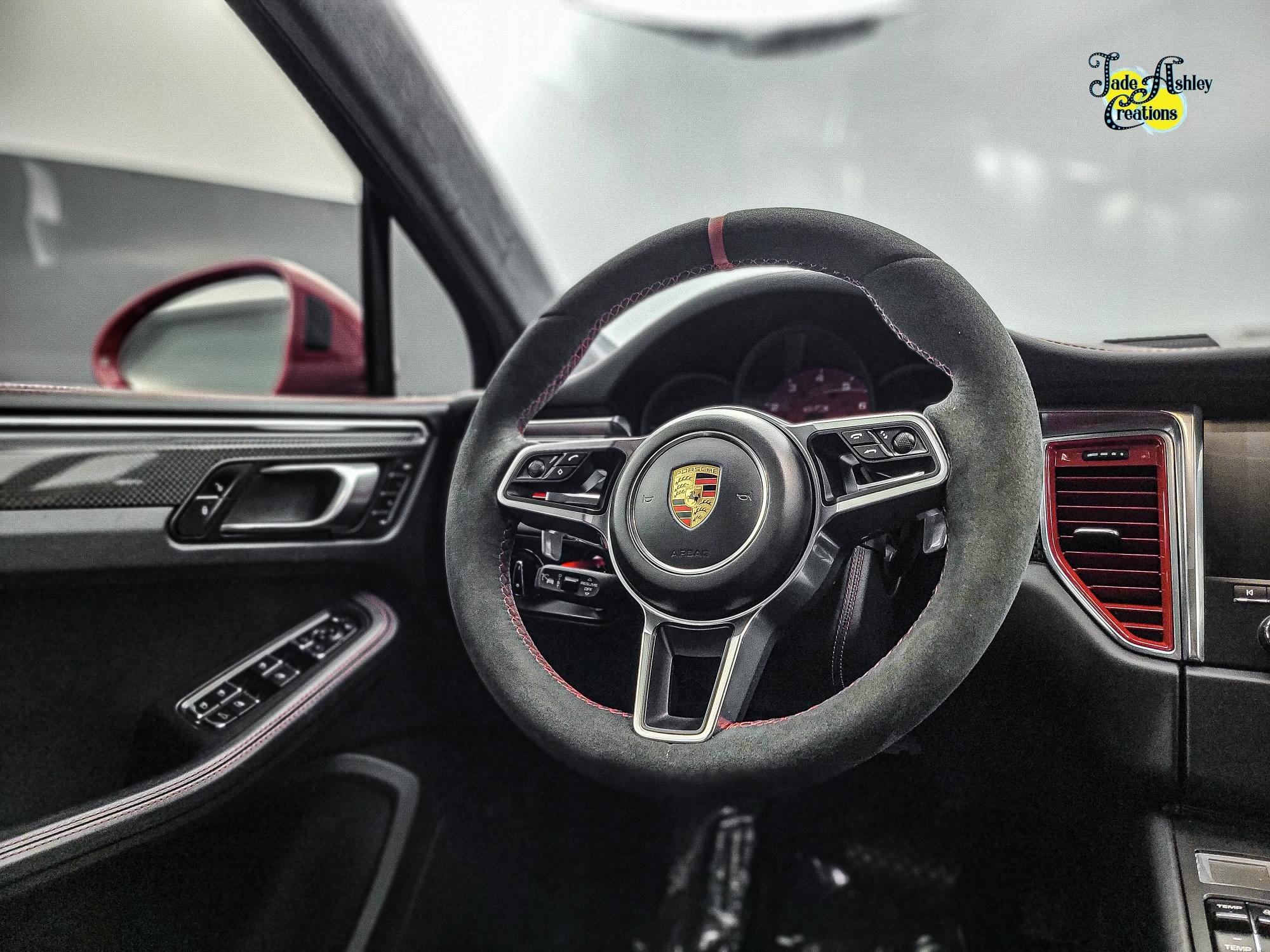 Interior of a Porsche car showcasing the steering wheel with Porsche emblem, side mirror, car dashboard, and red air vent.