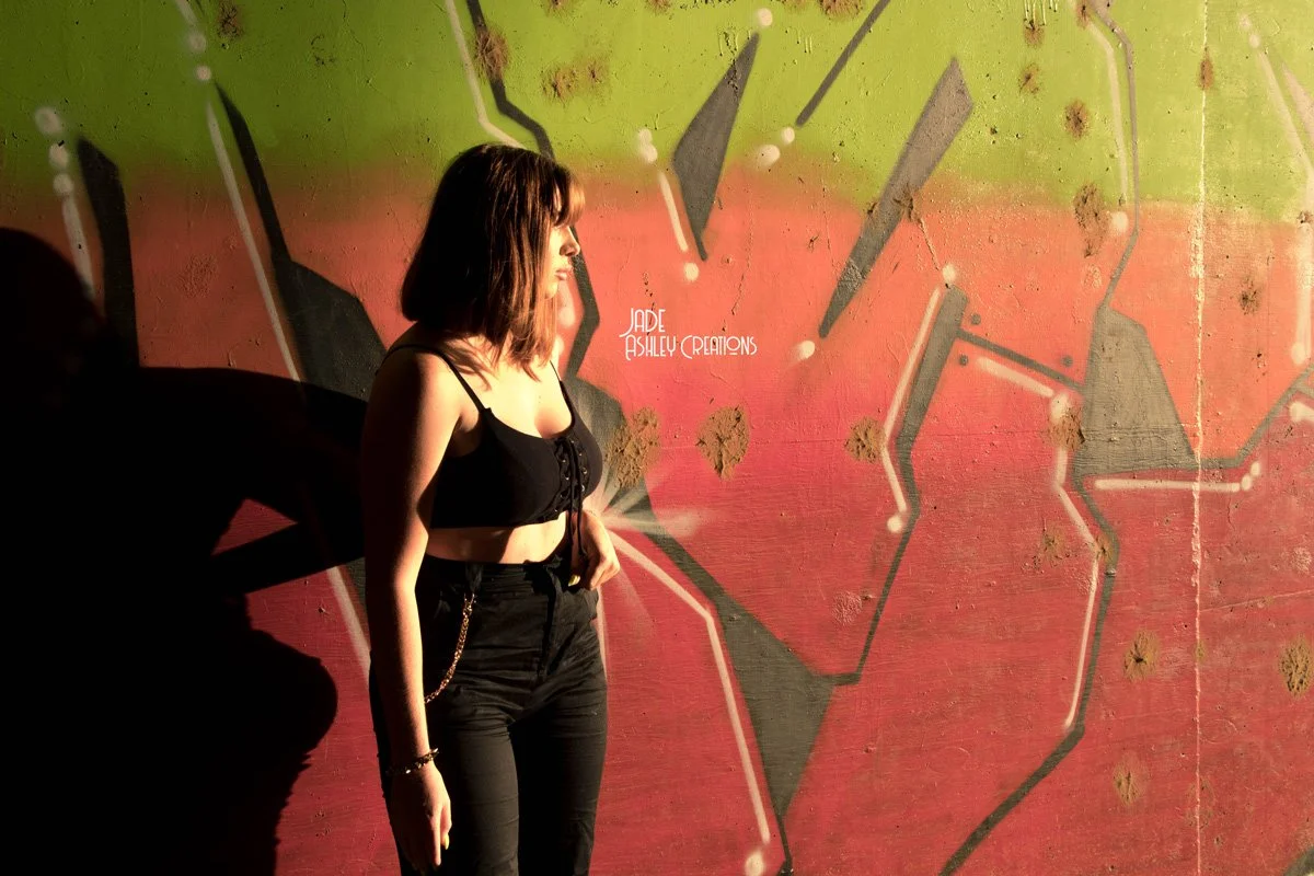 A woman with shoulder-length hair wearing a black crop top and black pants stands in front of a colorful graffiti wall.