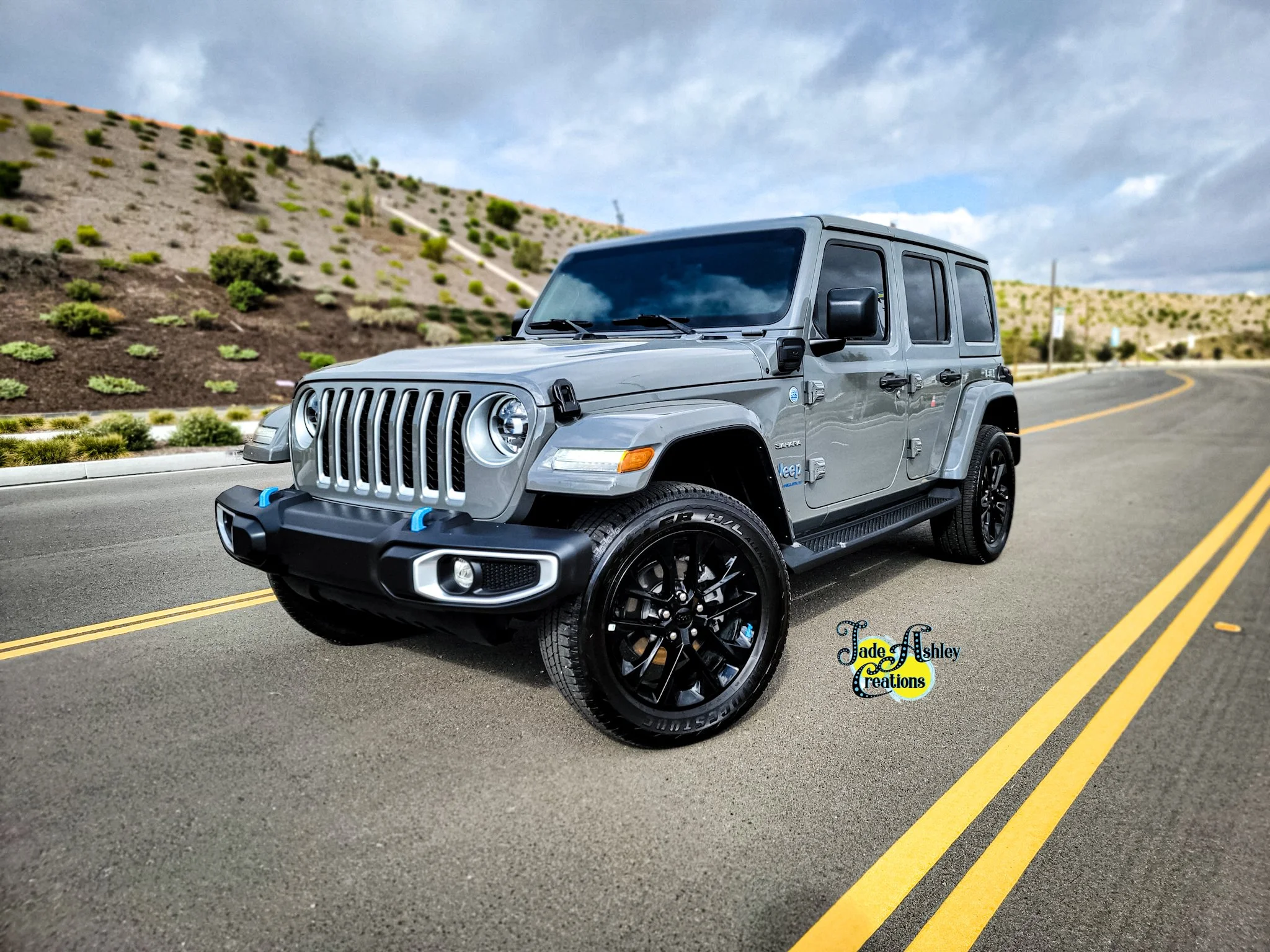 A gray Jeep Wrangler parked on a curved road with a hillside of small green shrubs in the background under a partly cloudy sky.