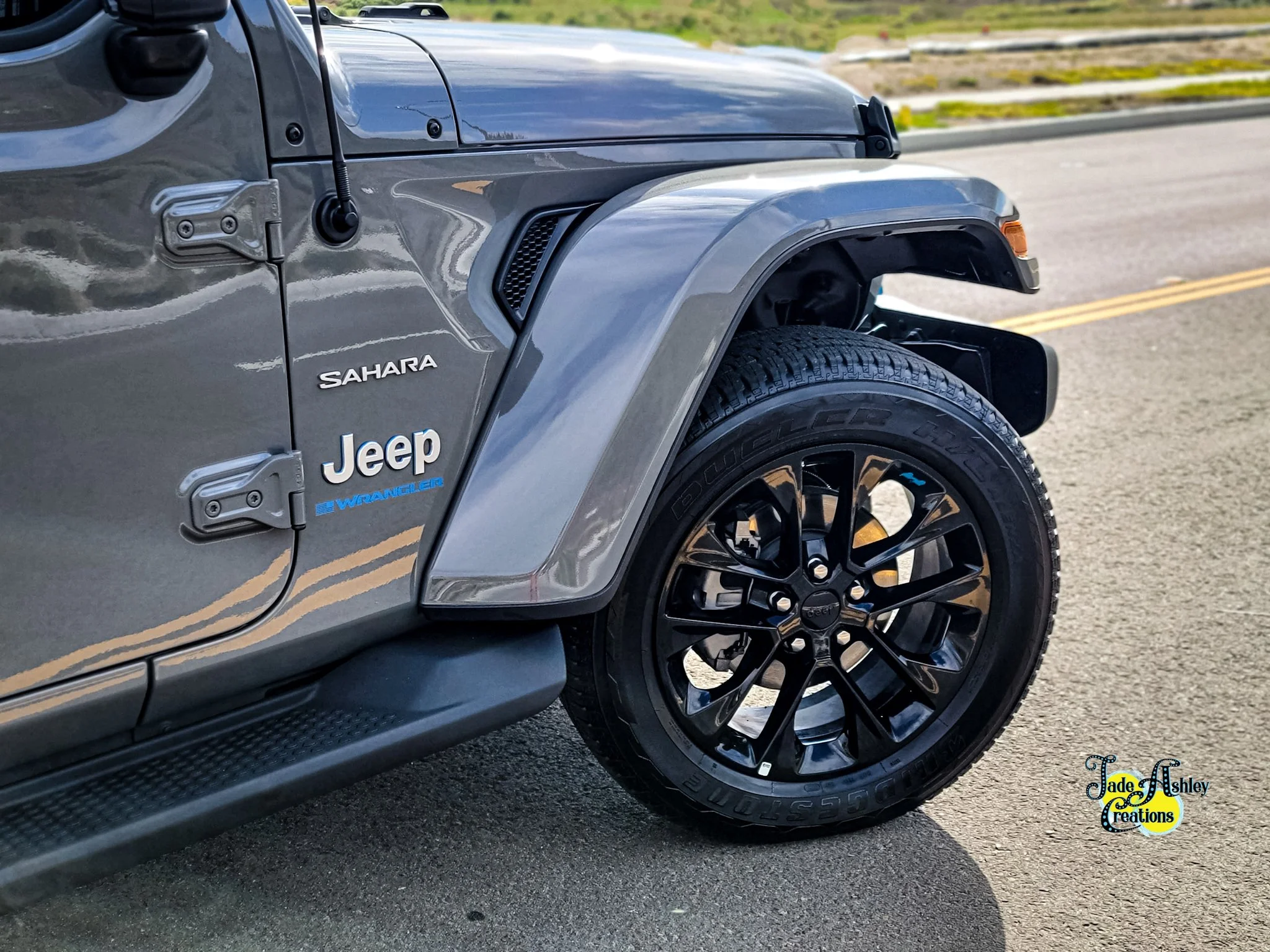 Close-up of the front left side of a gray Jeep Sahara Wrangler, showing the tire, wheel, fender, and part of the door with Jeep and Sahara badges.