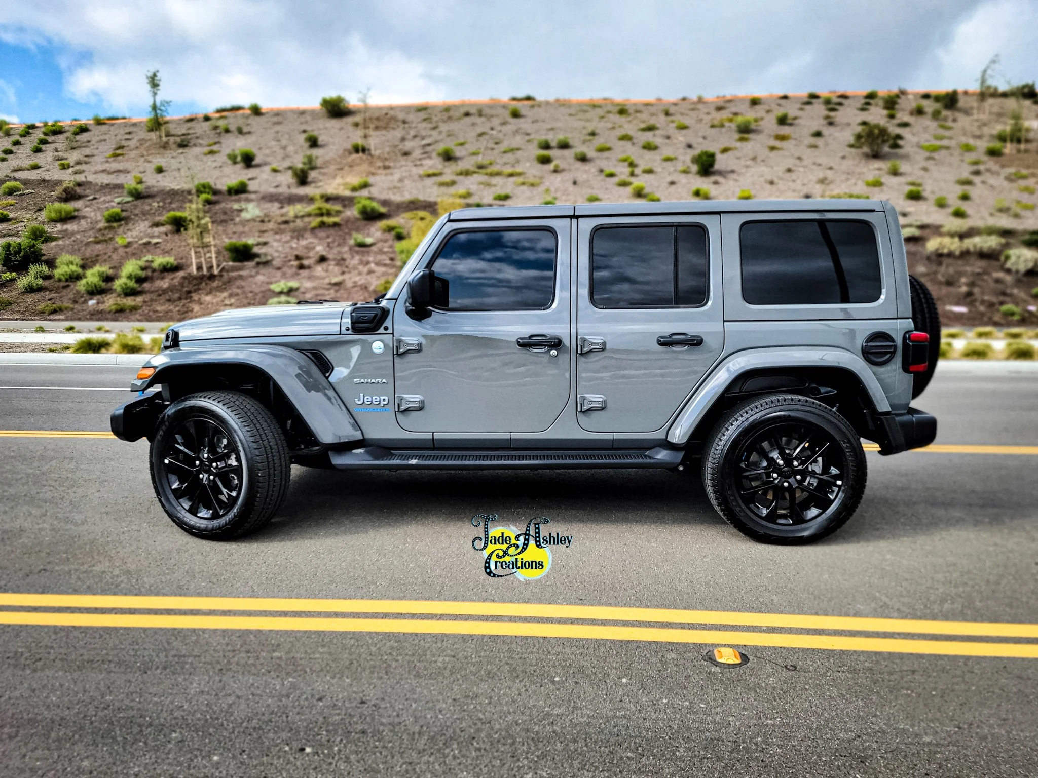 Gray Jeep Wrangler parked on a paved road with double yellow lines, a hillside with sparse greenery in the background, and partly cloudy sky.