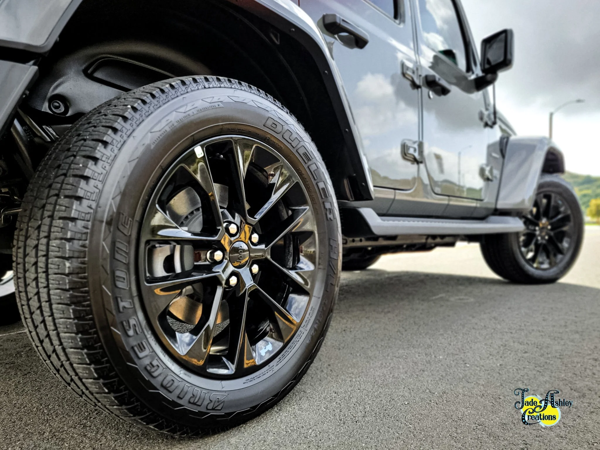 Close-up of a black modern SUV's front wheel and tire, with the vehicle parked on a paved surface under a partly cloudy sky, and a green landscape in the background.