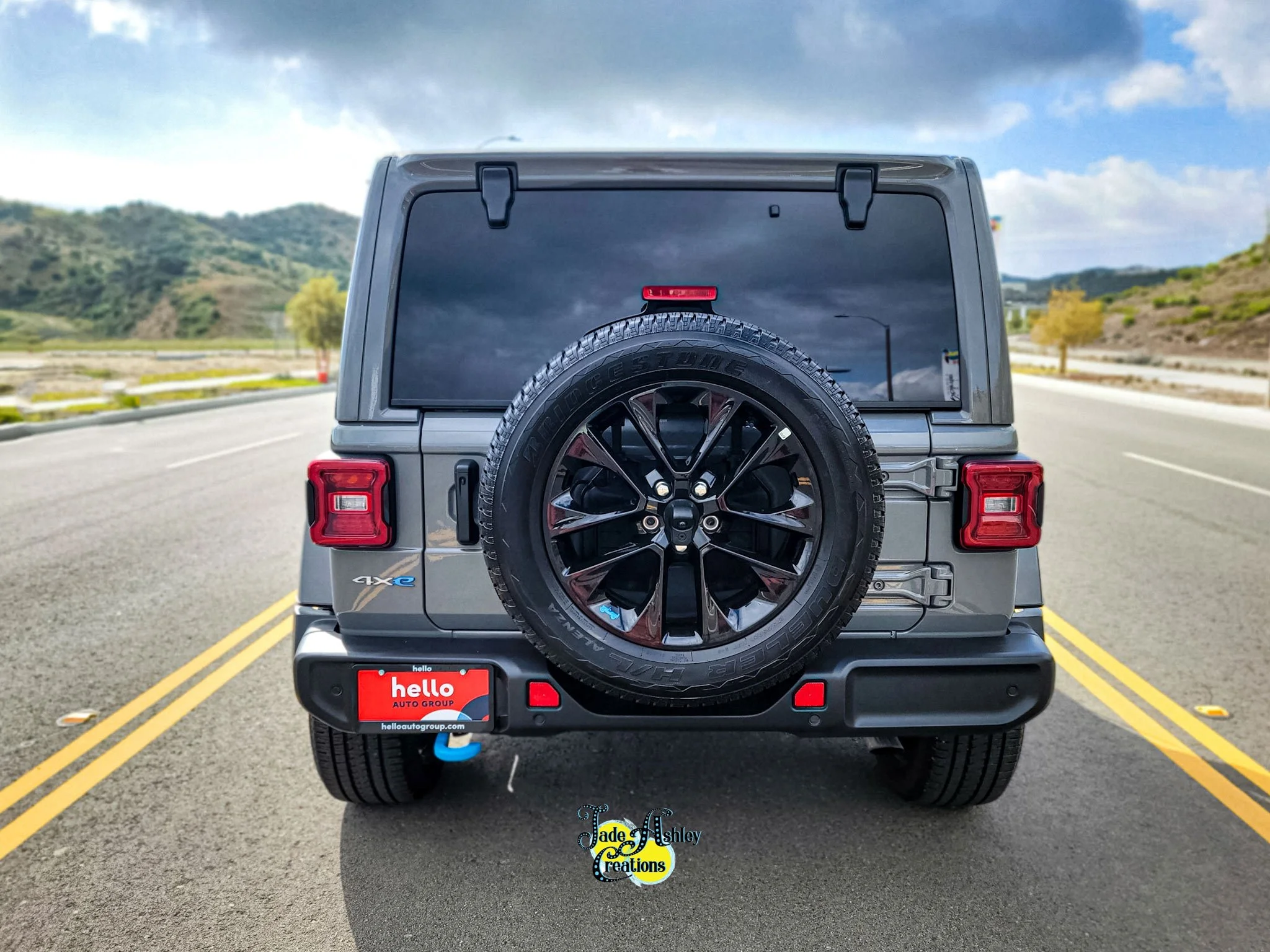 Rear view of a gray Jeep Wrangler with a spare tire mounted on the back, parked on an empty road with yellow double lines. The landscape features hills and trees, with a cloudy sky overhead.