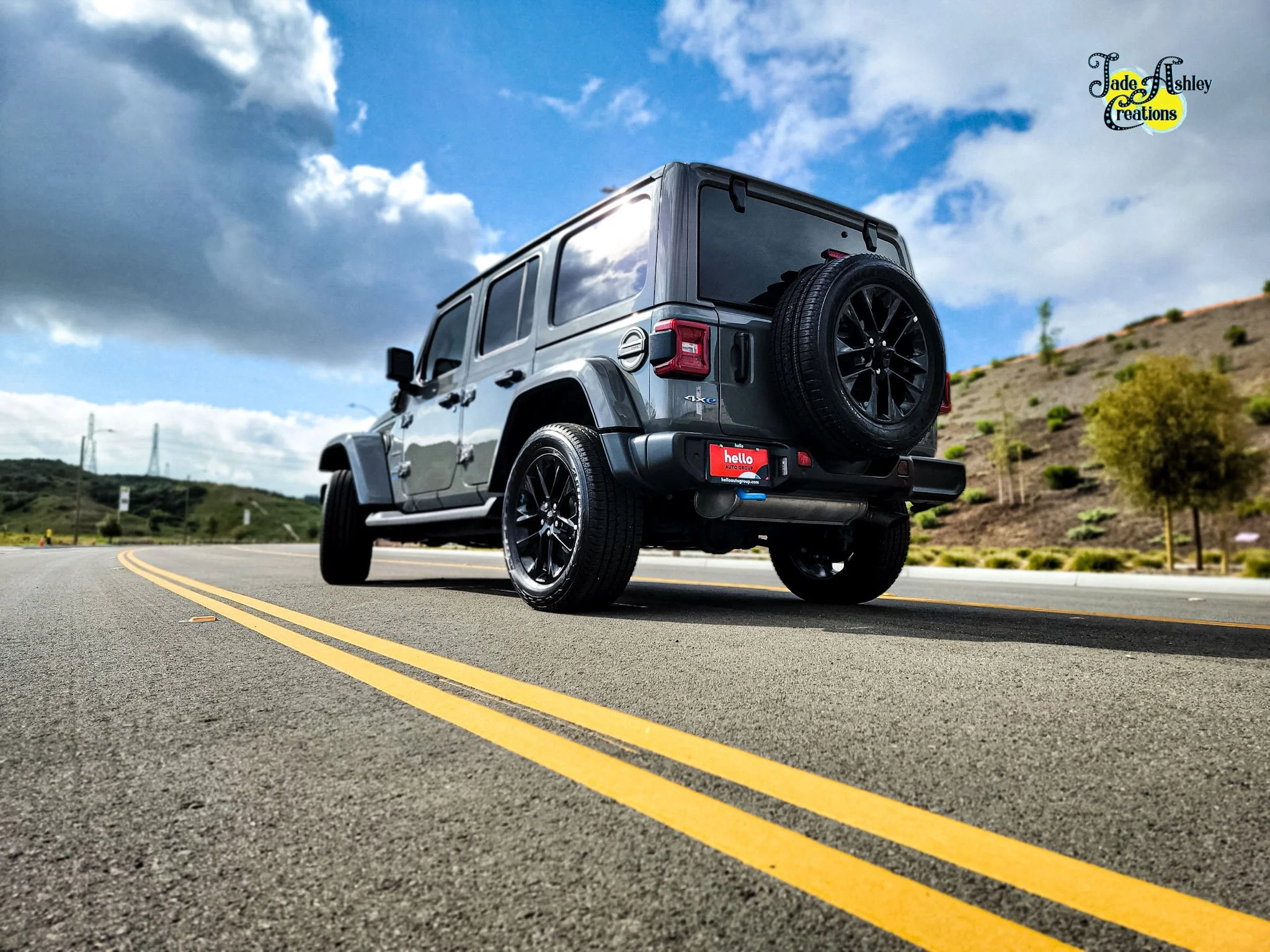 A gray Jeep SUV driving on a winding two-lane road in a hilly area with sparse trees, under a partly cloudy sky.