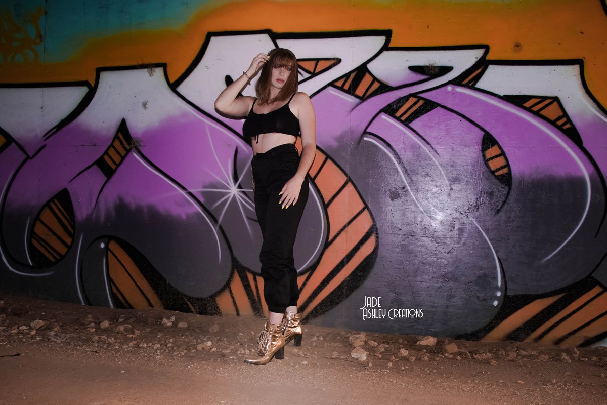 A woman with shoulder-length light brown hair, wearing a black crop top, black pants, and gold high-heeled boots, stands in front of a colorful graffiti mural on a wall. The mural features large, stylized letters with purple, orange, and black colors