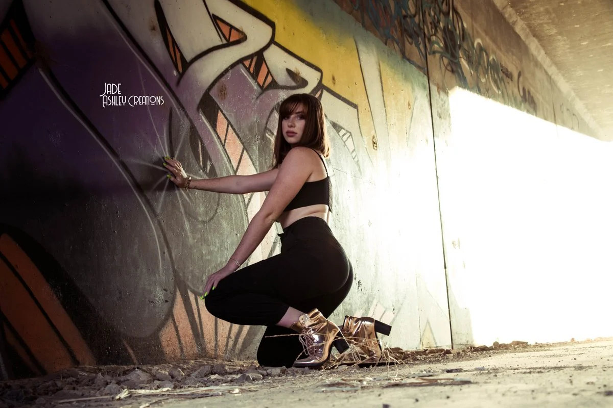 A woman wearing a black crop top, black pants, and gold high-heeled boots crouches against a graffiti-covered wall in an underpass.