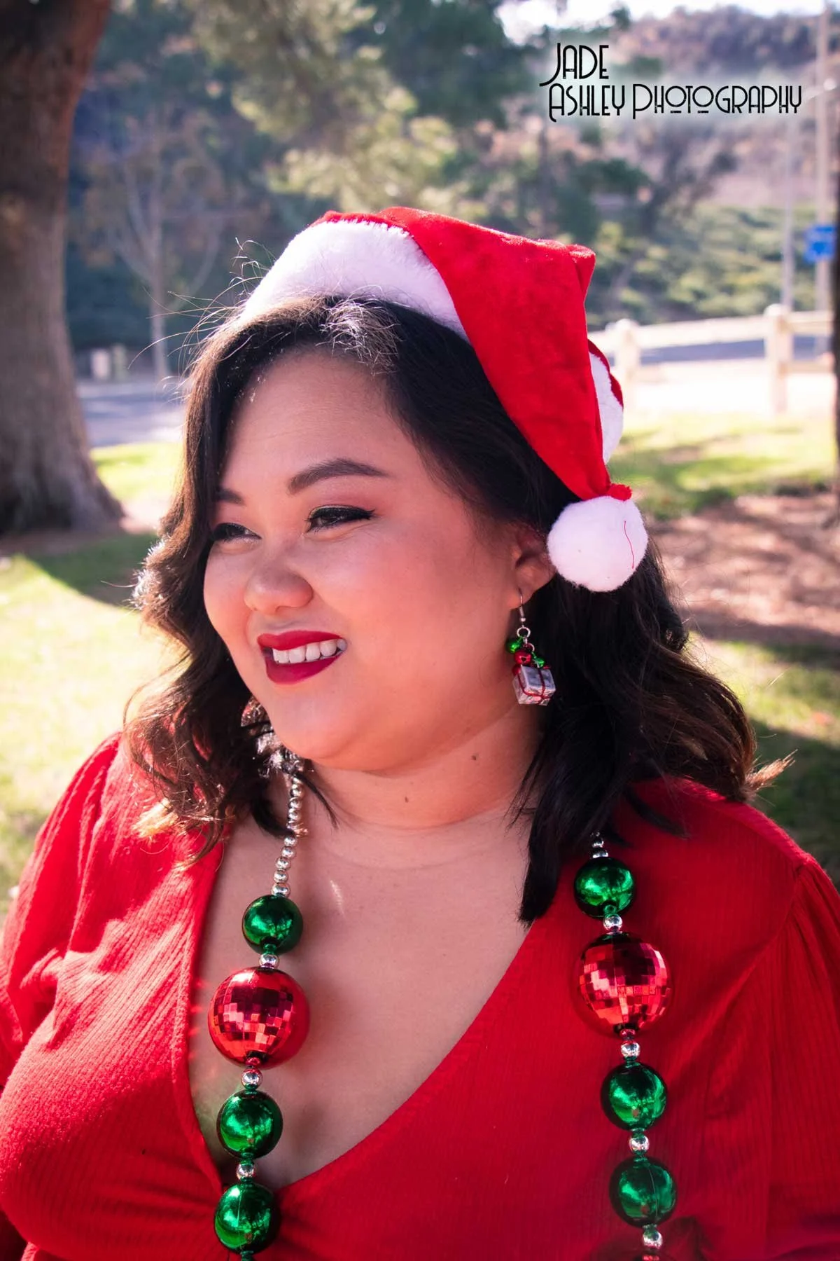 A woman wearing a Santa hat and Christmas-themed jewelry, smiling outdoors on a sunny day. She is dressed in red and has a Christmas necklace with large green and red beads.