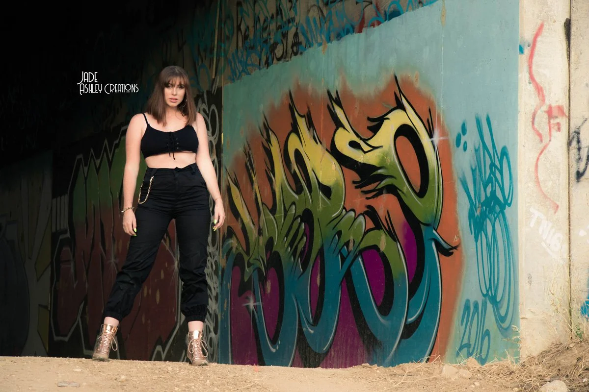 A young woman standing in front of a graffiti mural on a concrete wall, wearing a black crop top, black pants, and gold shoes.