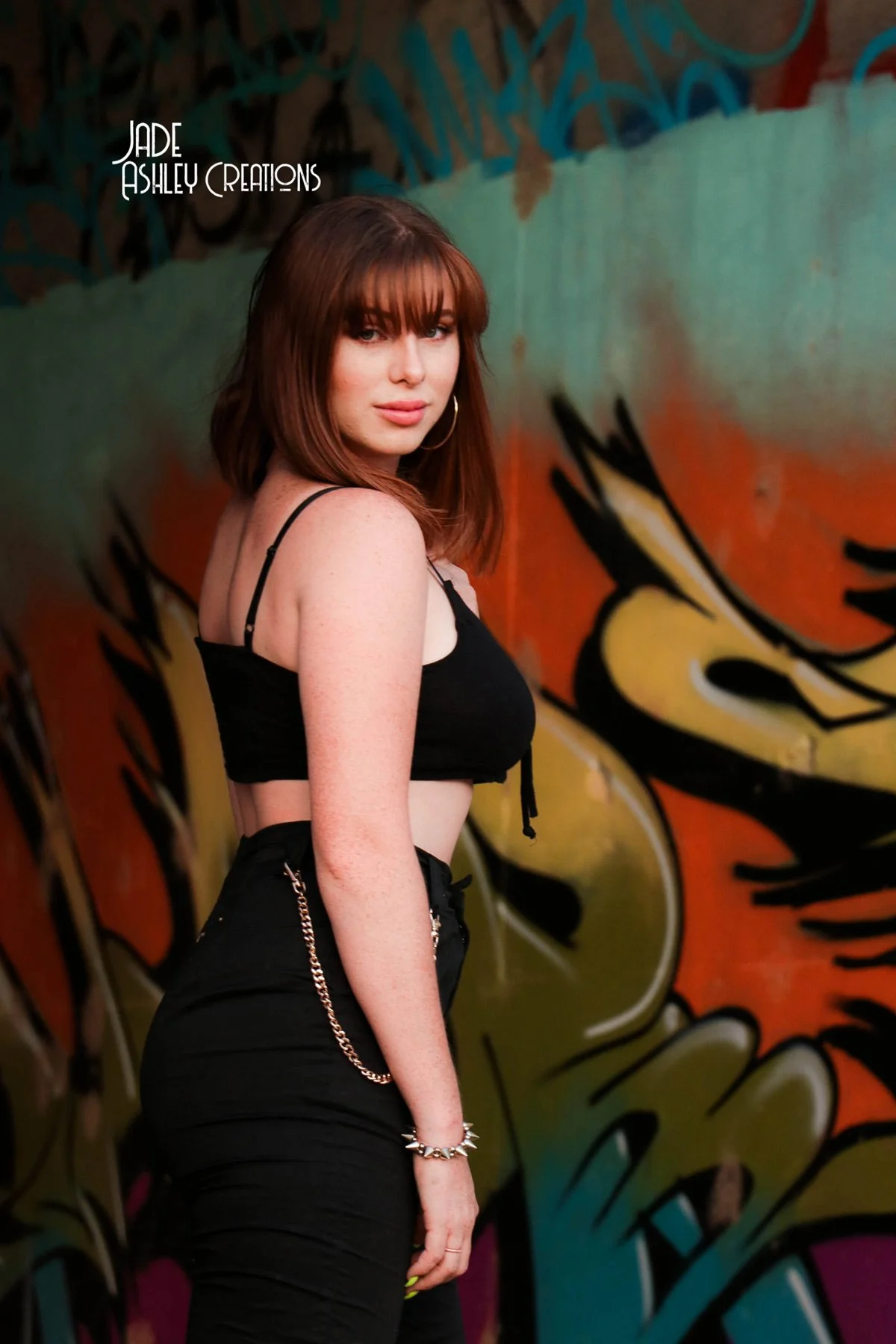 Young woman with reddish-brown hair and hoop earrings standing in front of a colorful graffiti wall, wearing a black crop top, black high-waisted pants, and chain jewelry.
