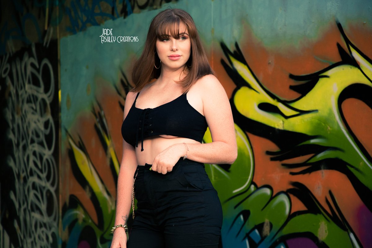 A young woman with brown hair and bangs stands in front of a colorful graffiti wall, wearing a black crop top and high-waisted black pants.