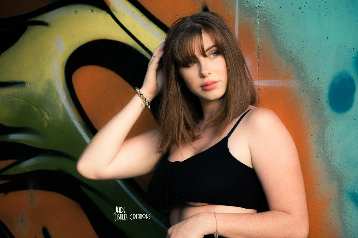 A woman with brown hair and light skin poses in front of a colorful graffiti wall, wearing a black crop top and jewelry.