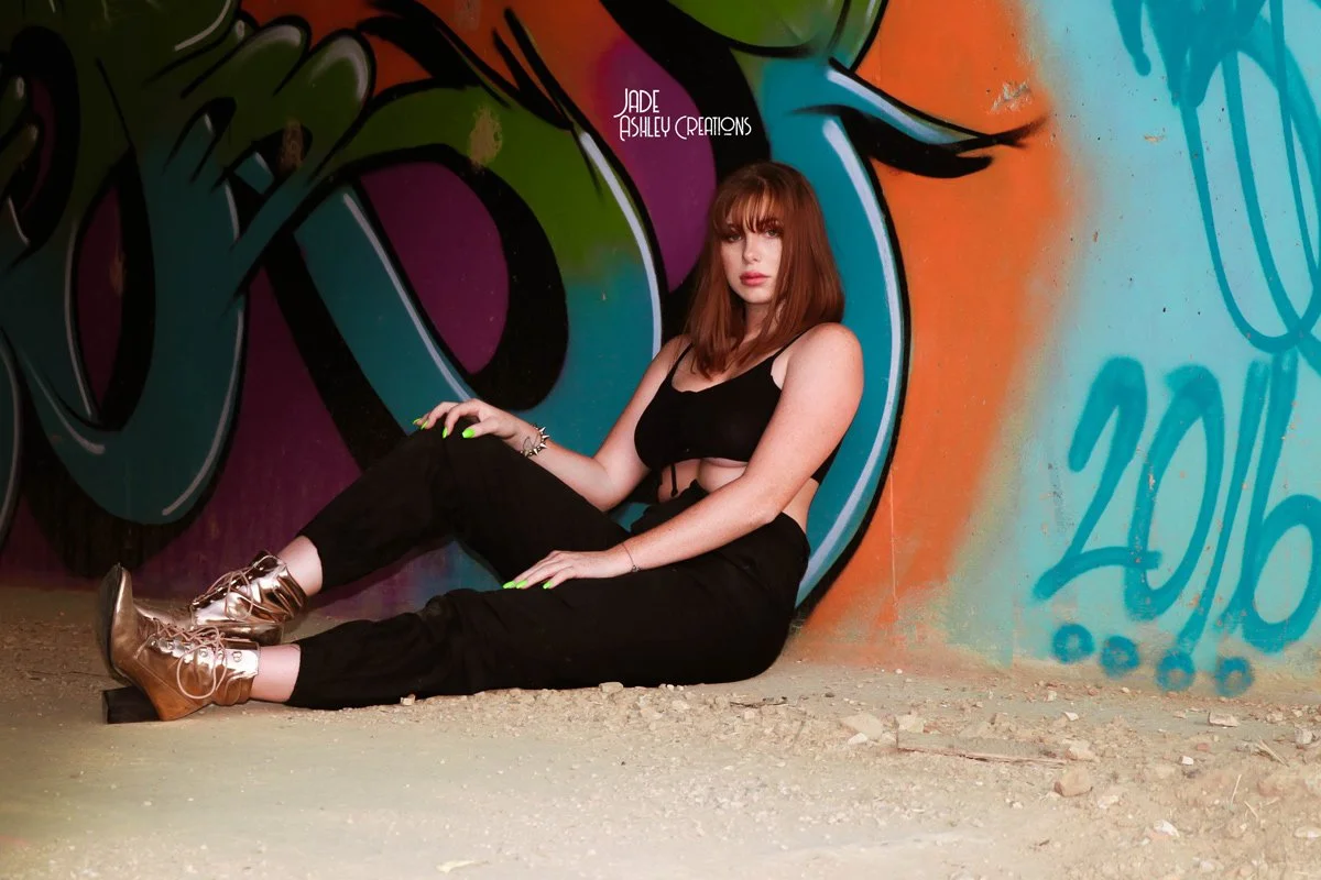 A woman with red hair, wearing a black crop top and black pants, sitting on the ground against a colorful graffiti wall with a large eye design and the year 2016 written on it.