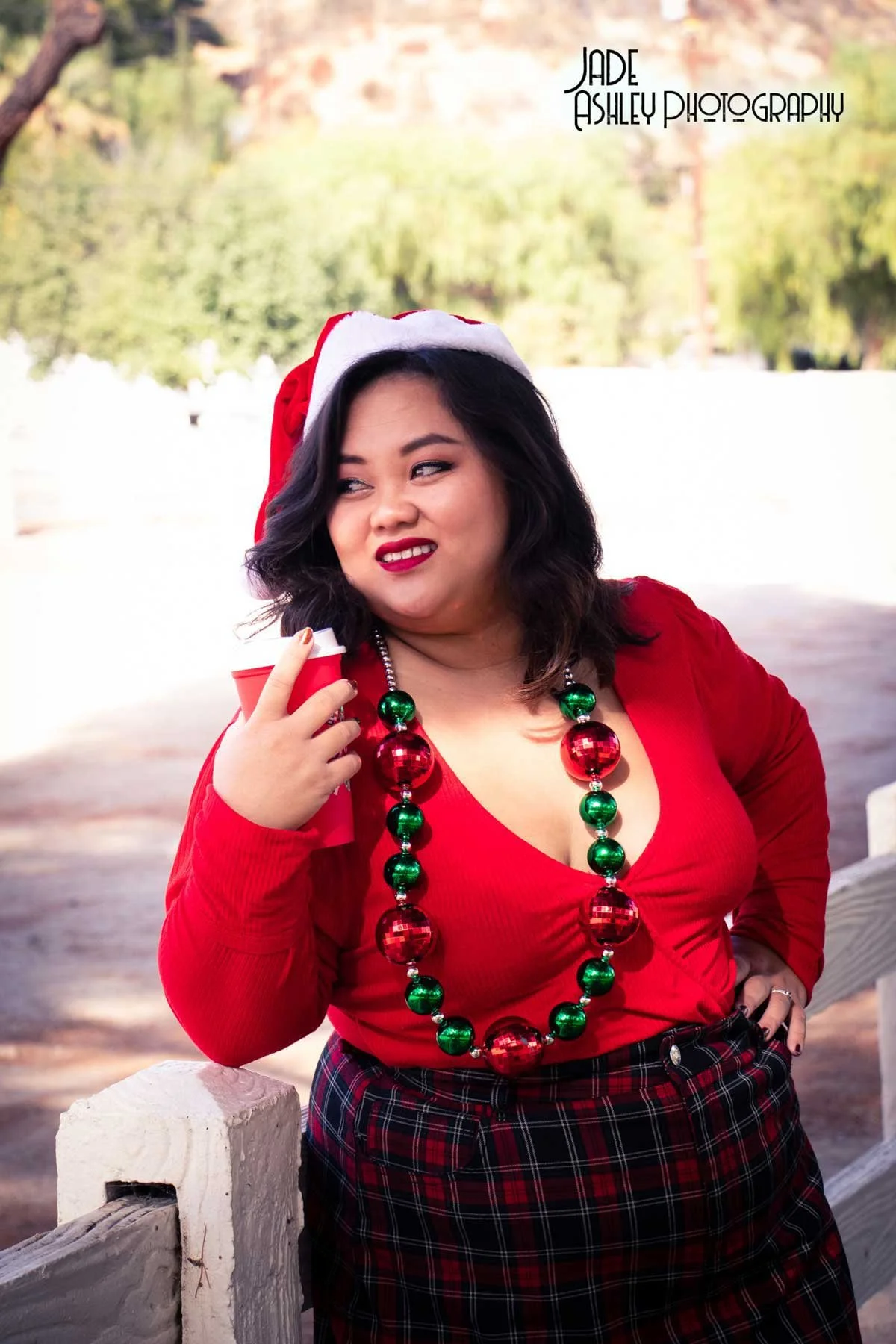 A woman wearing a red Santa hat and red top with a large red and green Christmas bead necklace, holding a red cup, standing outdoors with trees in the background.